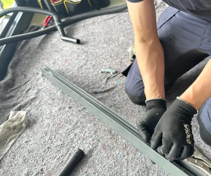 Monteur met zwarte werkhandschoenen knielt op een stucloper bij een losgenomen metalen railprofiel, met een tang op het profiel en op de achtergrond een stofzuigerslang en een gereedschapstas